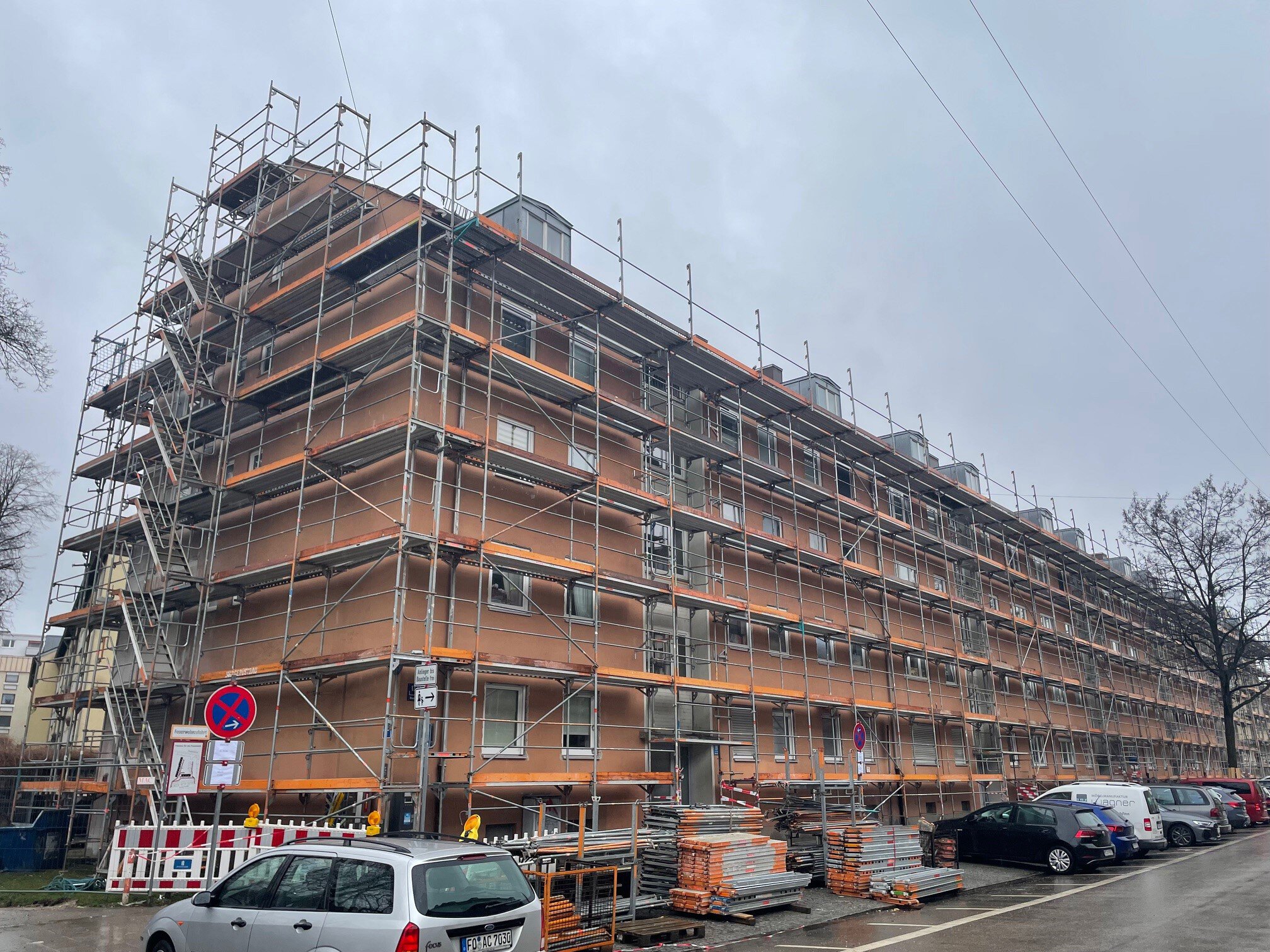 Weitwinkelaufnahme eines langen, mehrstöckigen Wohnblocks, dessen gesamte Fassade und Giebelseite vollständig eingerüstet sind; im Vordergrund stehen Baumaterialien und geparkte Autos an einer Straße unter grauem Himmel.