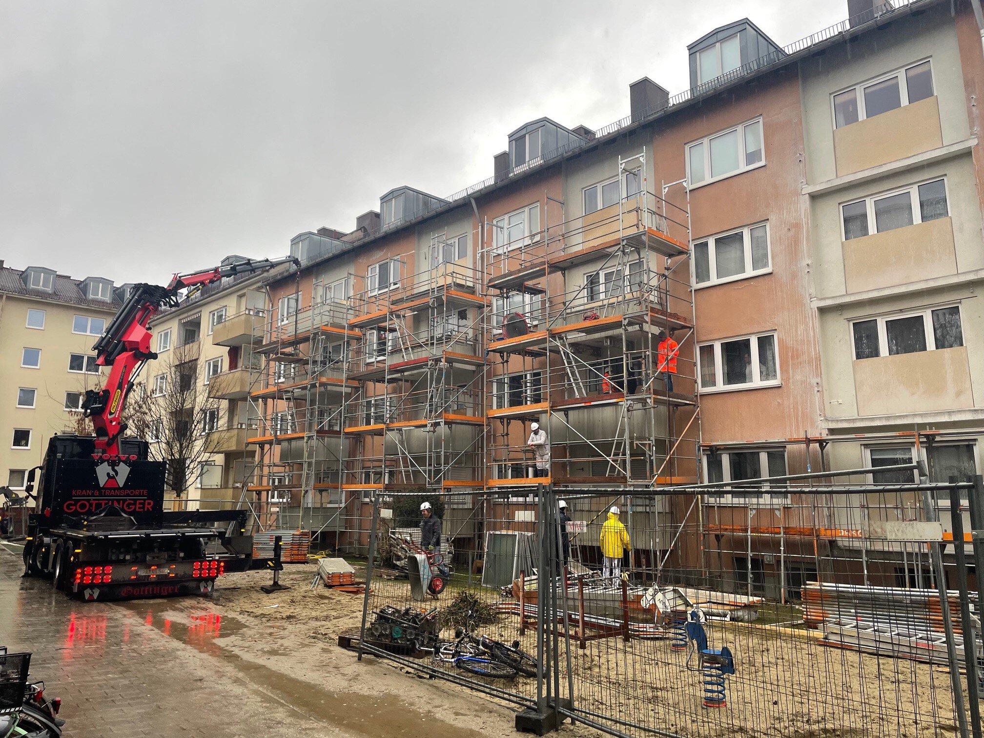 Eine Baustelle vor dem mehrstöckigen Wohnhaus des Bonner Quartiers mit einem großen roten Kran-LKW im Vordergrund und einem Metallgerüst an der Fassade, an dem Bauarbeiter bei bewölktem Himmel arbeiten.