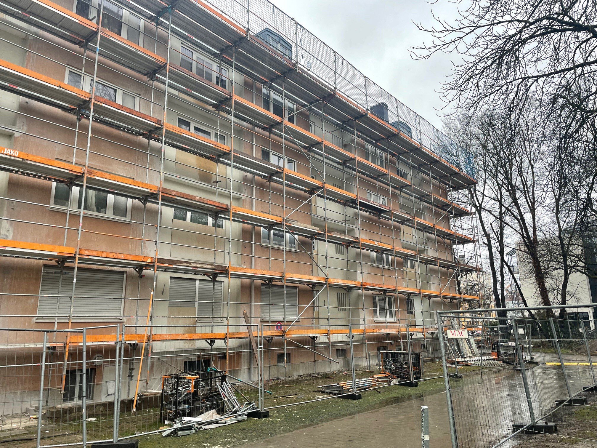 Die gesamte mehrstöckige Fassade eines Wohnhauses ist mit einem Baugerüst und Sicherheitsnetzen überzogen, davor steht ein Bauzaun auf einem asphaltierten Weg.