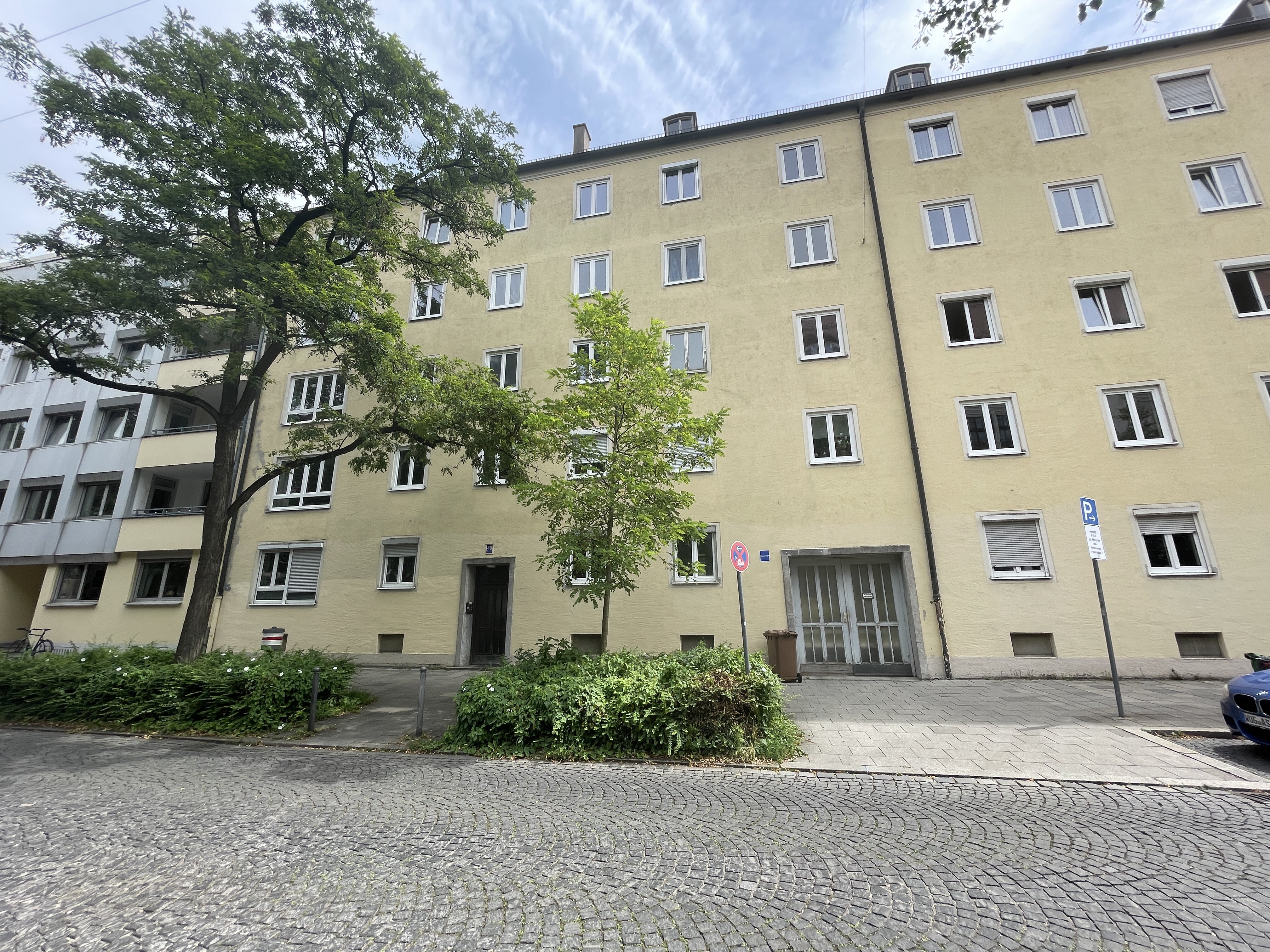 Mehrstöckiges Wohngebäude der Hiltensbergerstraße in München mit gelber Fassade, mehreren Fenstern und zwei Eingängen. Im Vordergrund befinden sich eine gepflasterte Straße, sowie ein Baum und Grünflächen. Der Himmel ist teilweise bewölkt