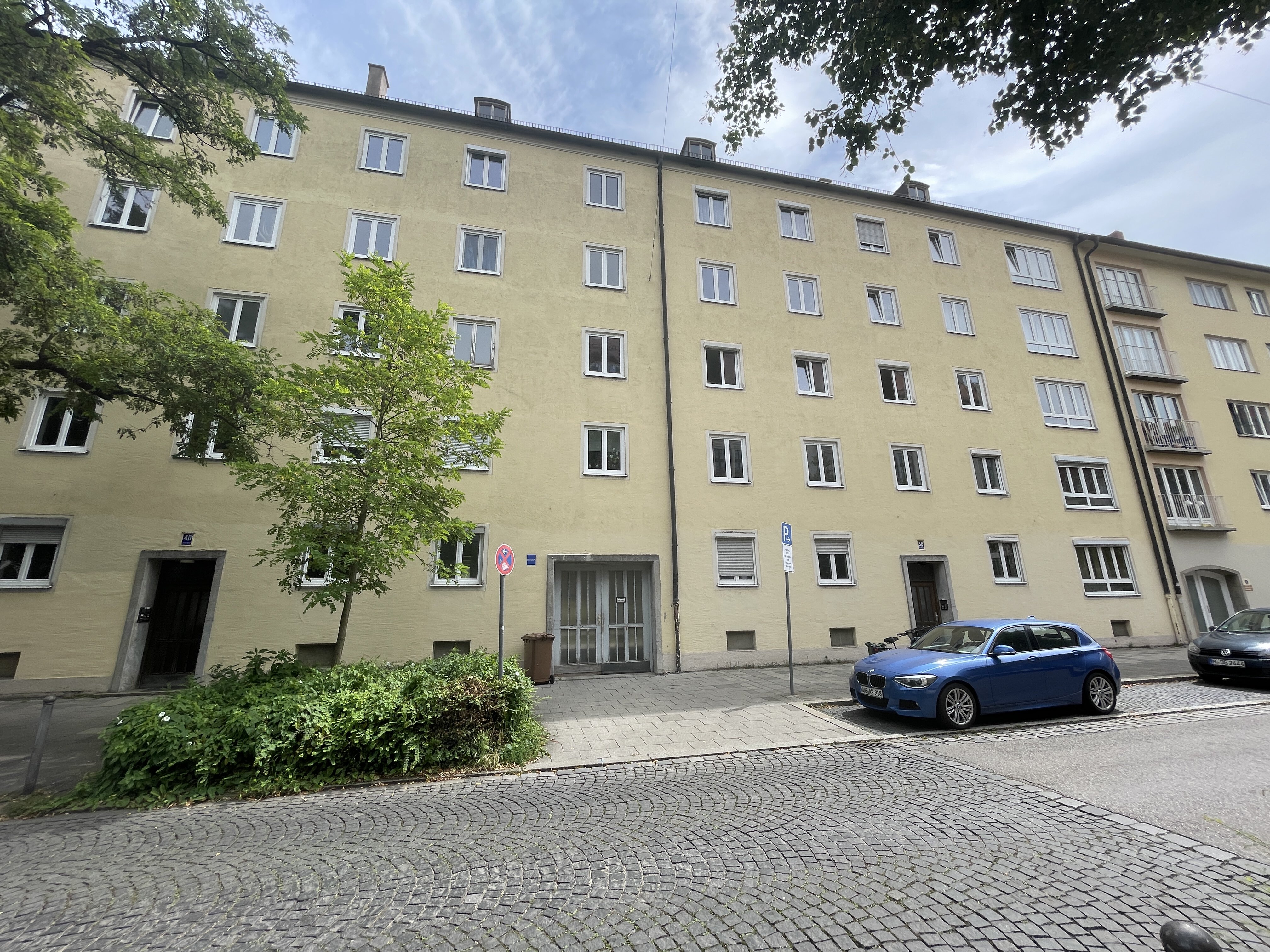Mehrstöckiges Wohngebäude der Hiltensbergerstraße in München mit gelber Fassade, mehreren Fenstern und zwei Eingängen. Im Vordergrund befinden sich eine gepflasterte Straße, parkende Autos sowie ein Baum und Grünflächen. Der Himmel ist teilweise bewölkt.
