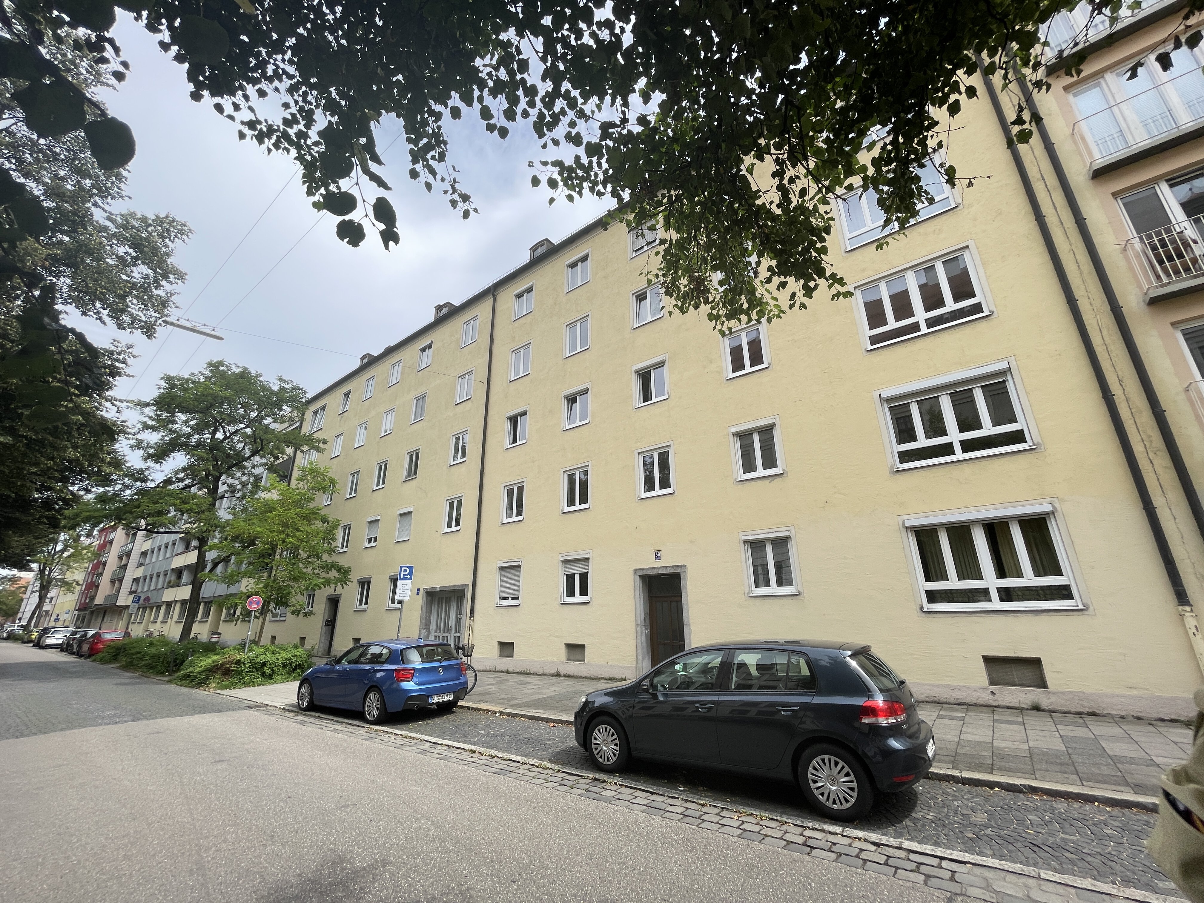 Hiltensbergerstraße in München mit mehrstöckigen Wohnhaus mit gelber Fassade, mehreren Fenstern und zwei Eingängen im Vordergrund. Neben den Häusern der gepflasterten Straße sieht man parkenden Autos sowie ein Baum und Grünflächen. Der Himmel ist teilweise bewölkt