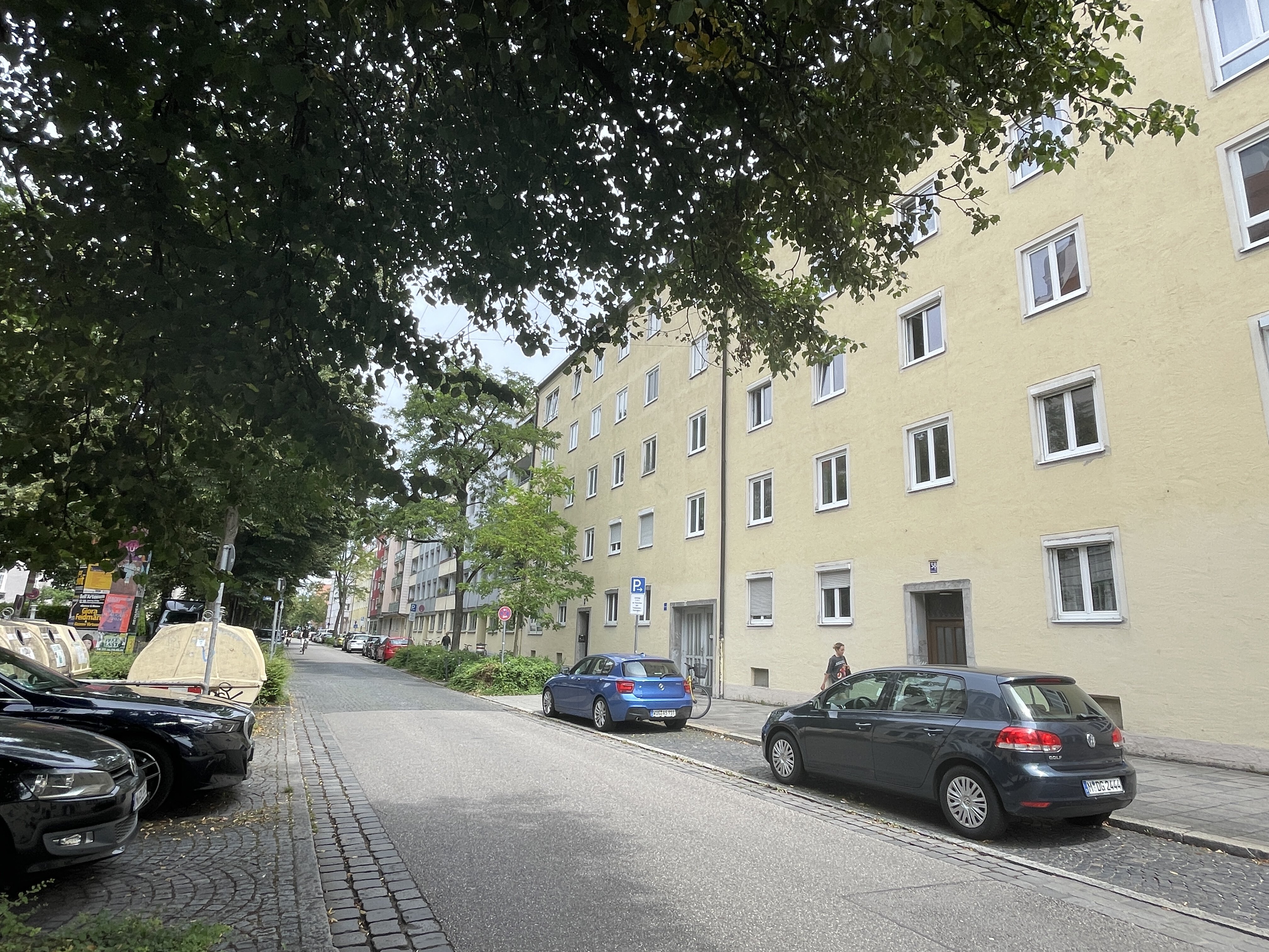 Hiltensbergerstraße in München mit mehrstöckigen Wohnhaus mit gelber Fassade, mehreren Fenstern und zwei Eingängen im Vordergrund. Neben den Häusern der gepflasterten Straße sieht man parkenden Autos sowie ein Baum und Grünflächen. Der Himmel ist teilweise bewölkt