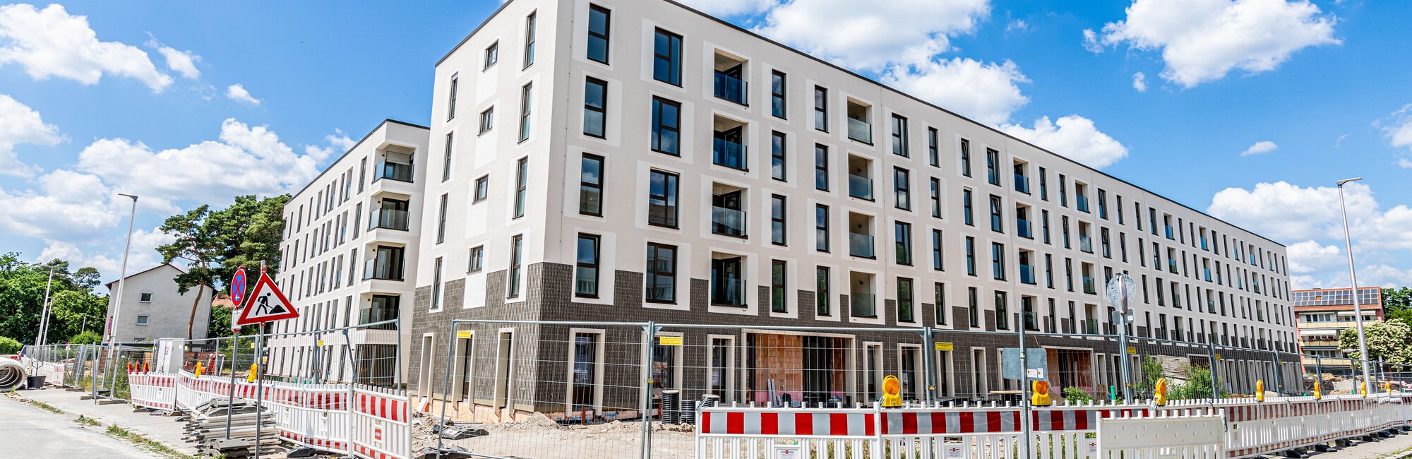 Ein langes, modernes, weißes Wohngebäude mit dunklen Fensterrahmen und einem dunklen Sockel ist im Bau und von rot-weißen Bauzäunen umgeben. Links und rechts führen Straßen entlang.