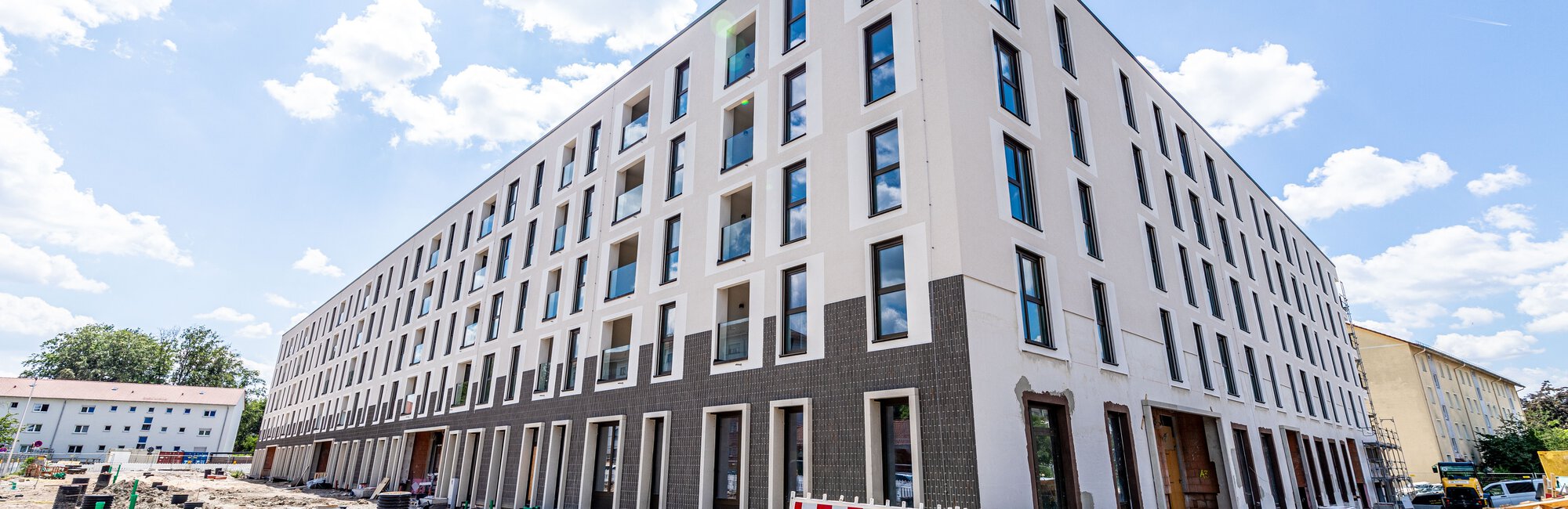  Ein langes, modernes, weißes Wohngebäude mit dunklen Fensterrahmen und einem dunklen Sockel ist im Bau und von rot-weißen Bauzäunen umgeben. Links und rechts führen Straßen entlang.
