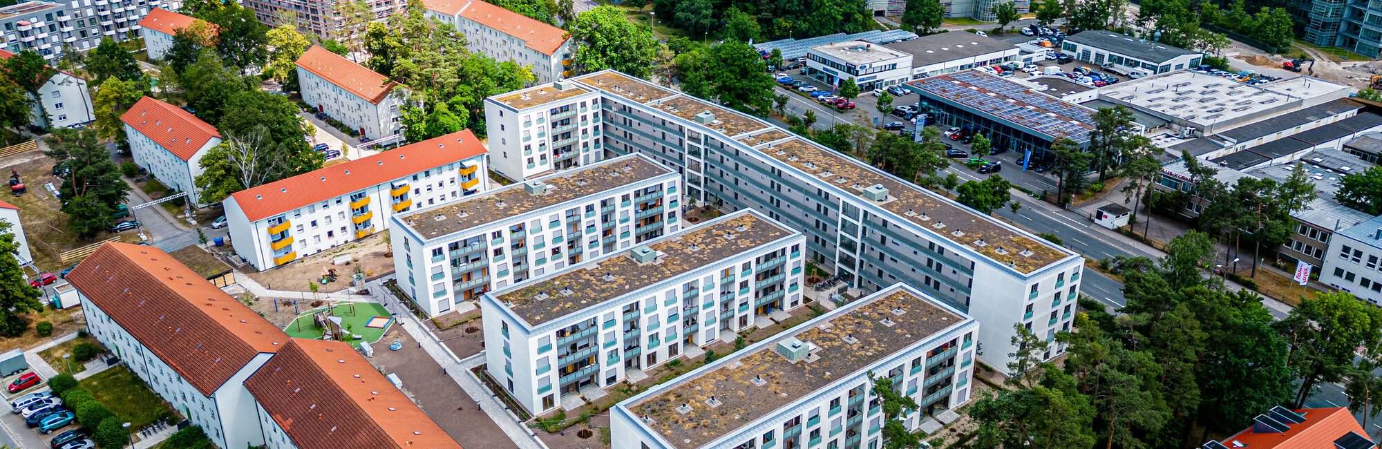  Luftaufnahme einer Wohnsiedlung mit mehreren modernen, weißen Mehrfamilienhäusern, die von älteren Gebäuden mit roten Dächern und viel Grün umgeben sind. Im Hintergrund ist ein größeres Stadtgebiet mit weiteren Gebäuden und Wäldern zu sehen.