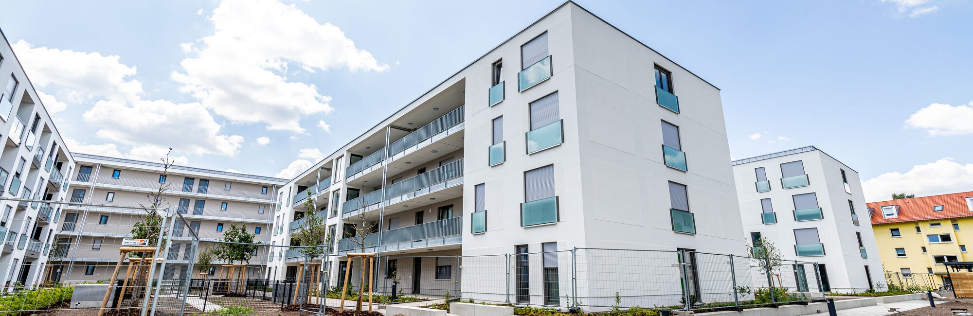 Ein moderner Gebäudekomplex mit weißen Fassaden und blauen Glasbalkonen unter einem bewölkten Himmel. Im Vordergrund befindet sich ein noch unbebauter Außenbereich mit Wegen und einigen jungen Pflanzen.