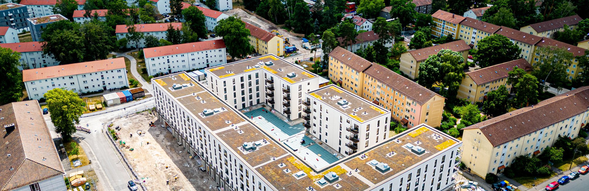  Luftaufnahme eines großen, modernen U-förmigen Wohnkomplexes mit einem Innenhof, umgeben von älteren Wohngebäuden und viel Grün. Davor und daneben befinden sich Straßen mit parkenden Autos.