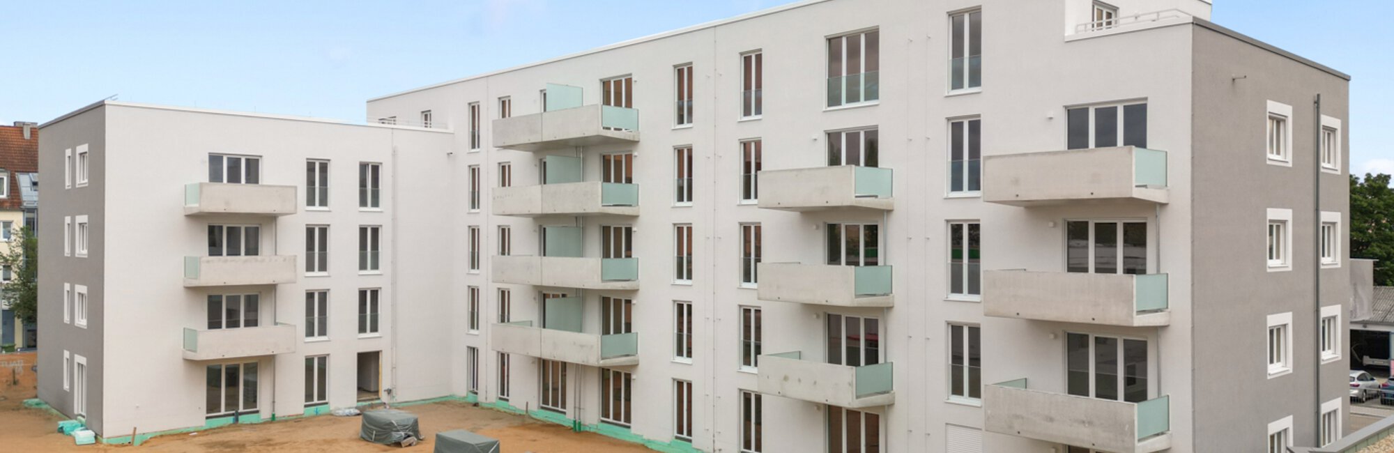 Neu gebautes, modernes Wohngebäude in Weiß und Grau mit Balkonen. Davor ein unbebauter Erdbereich, rechts unten ein Teil eines Gründaches.