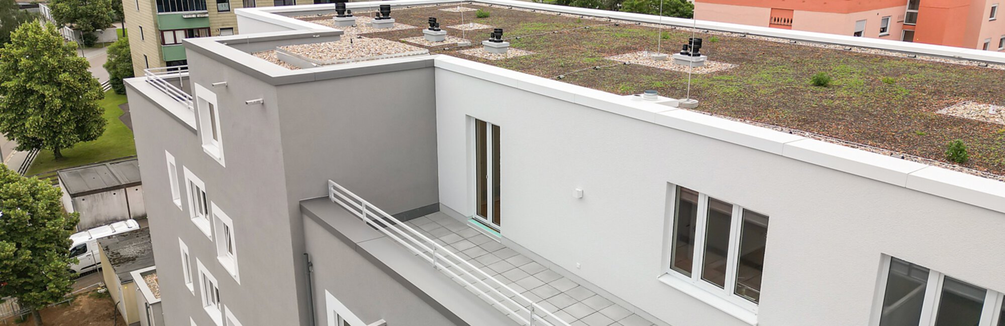 Modernes graues Wohngebäude mit Gründach und Dachterrasse. Im Hintergrund weitere Wohnblöcke und Landschaft.