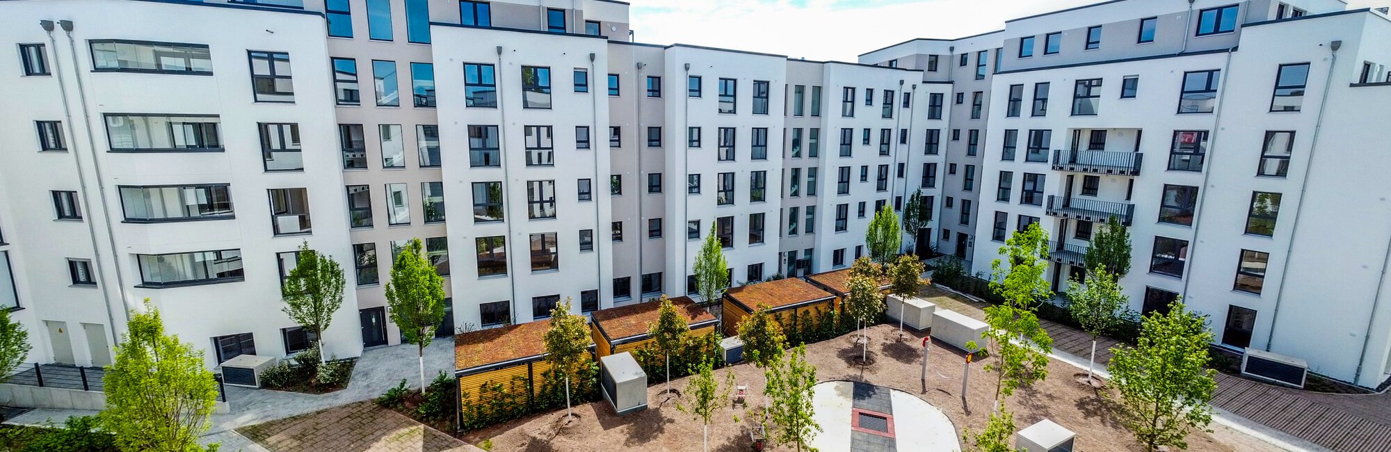  Ein moderner, weißer U-förmiger Wohnkomplex mit vielen Fenstern umschließt einen begrünten Innenhof mit Bäumen, Sitzgelegenheiten und gepflasterten Wegen. Im Vordergrund ist ein Teil eines weiteren Weges und junger Bepflanzung zu sehen. Das Datum "16.05.2022" ist unten links eingeblendet.