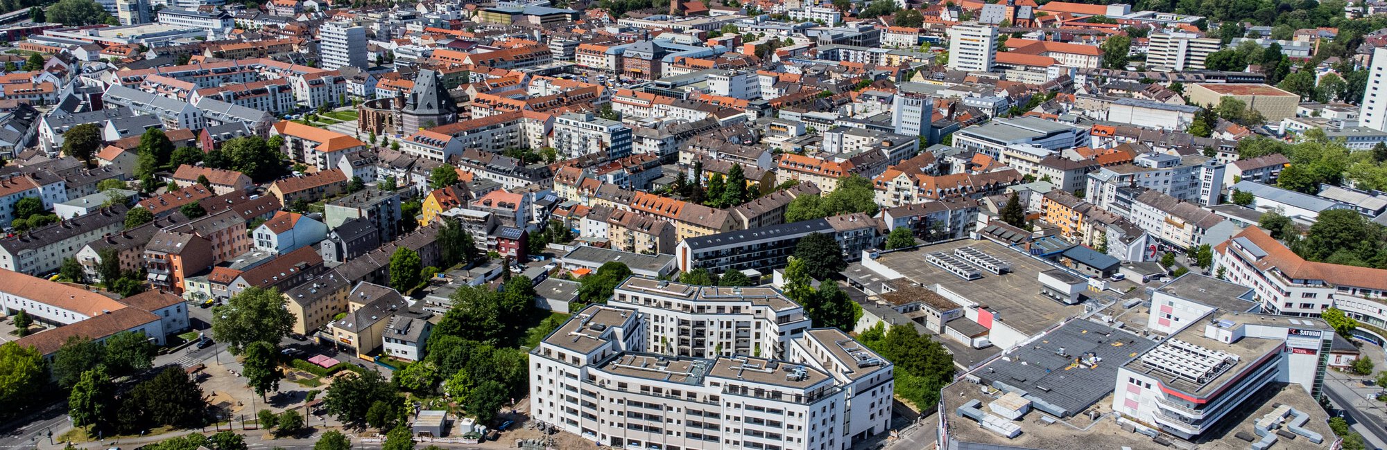 Luftaufnahme einer weitläufigen Stadtlandschaft mit vielen Gebäuden unterschiedlicher Größe und Dächerfarben, darunter ein großer, weißer Gebäudekomplex im Vordergrund. Die Stadt ist von Grünflächen und Wäldern umgeben. Das Datum "16.05.2022" ist unten links eingeblendet.