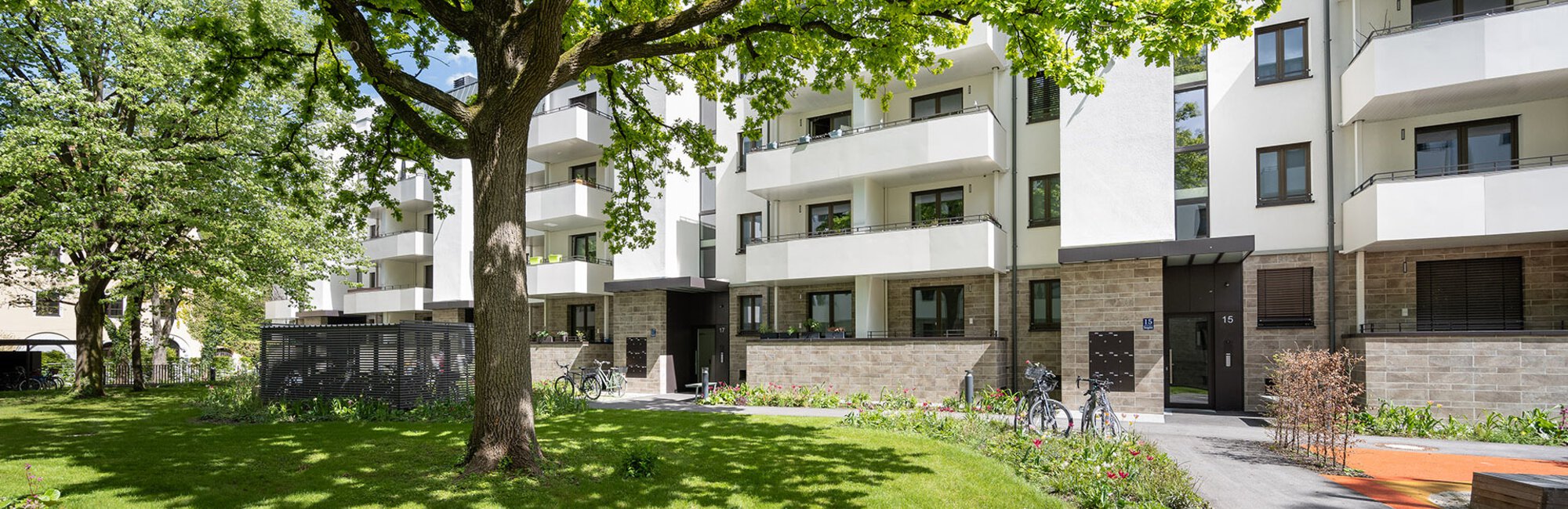 Blick von grüner Wiese auf modernes Wohngebäude mit weißer Fassade, Erdgeschoss mit Steinvertäfelung und geräumigen Balkonen auf jeder Etage. 